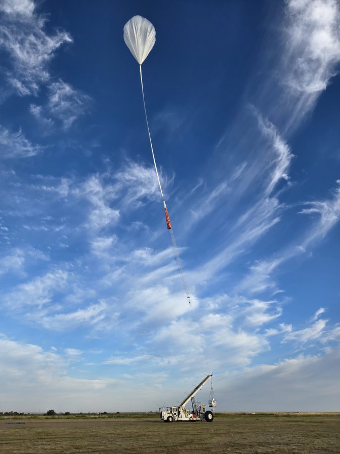 A large white scientific balloon in the shape of an upside down tear drop floats high in a bright blue sky with wispy white clouds, suspended by a long tether line connected to a white vehicle with a crane visible on the ground below in an open field. The crane is holding the BOOP experiment.