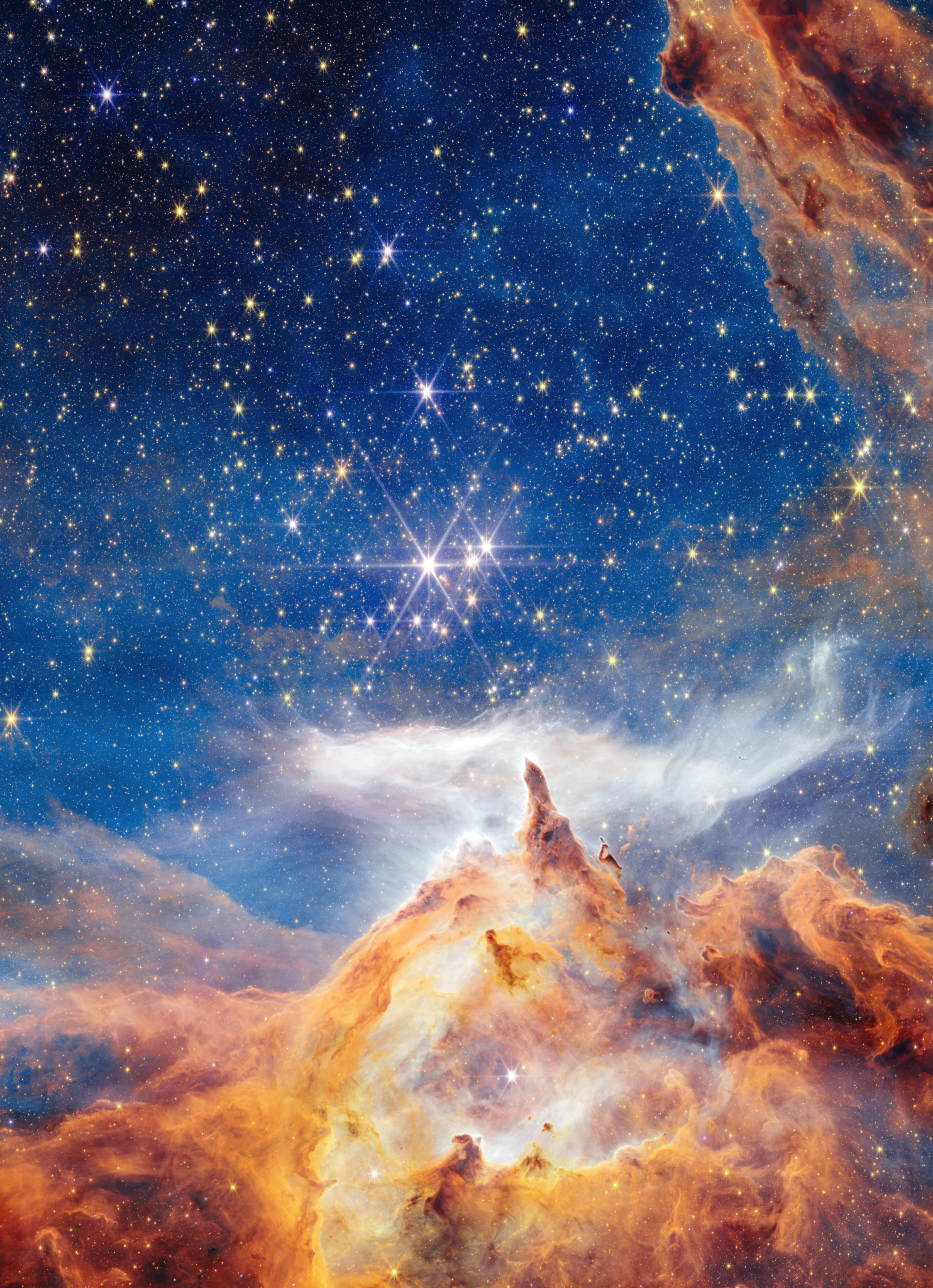 In what appears as a celestial dreamscape, a blue and black sky filled with brilliant stars covers about two thirds of the image. The stars are different sizes and shades of white, beige, yellow, and light orange. Across the bottom third of the scene is a craggy, mountain-like vista with spire-like peaks and deep, seemingly misty valleys. These so-called mountains appear in varying shades of orange, yellow, and brown. The soaring spires are going up, up, up, where a wispy, ethereal white cloud stretches horizontally across the scene. Steam appears to rise from the mountaintops and join with this cloud. At the top, right corner of the image, a swath of orange and brown structure cuts diagonally across the sky.