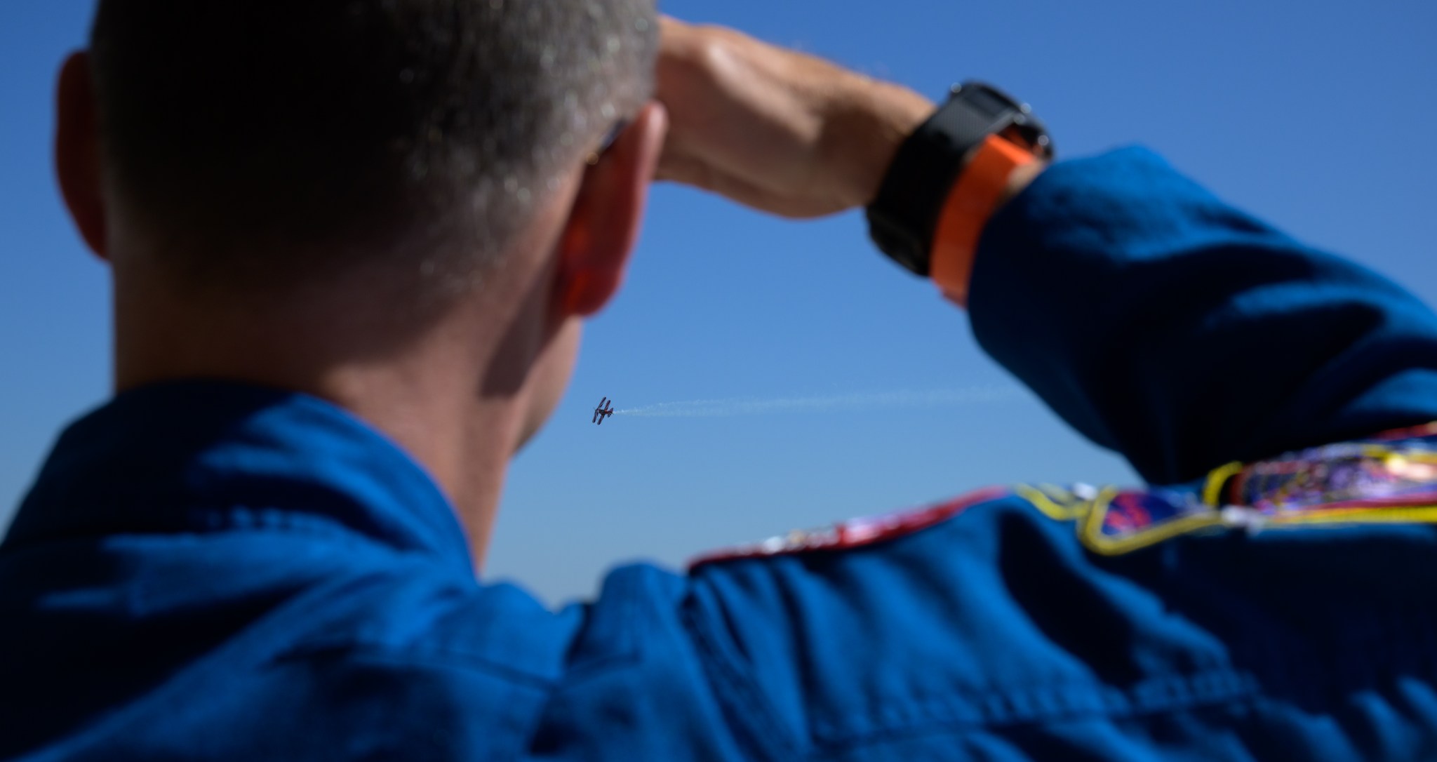 A man in a dark blue jumpsuit faces away from the camera, watching a plane in the light blue sky. He shades his eyes with his right hand.