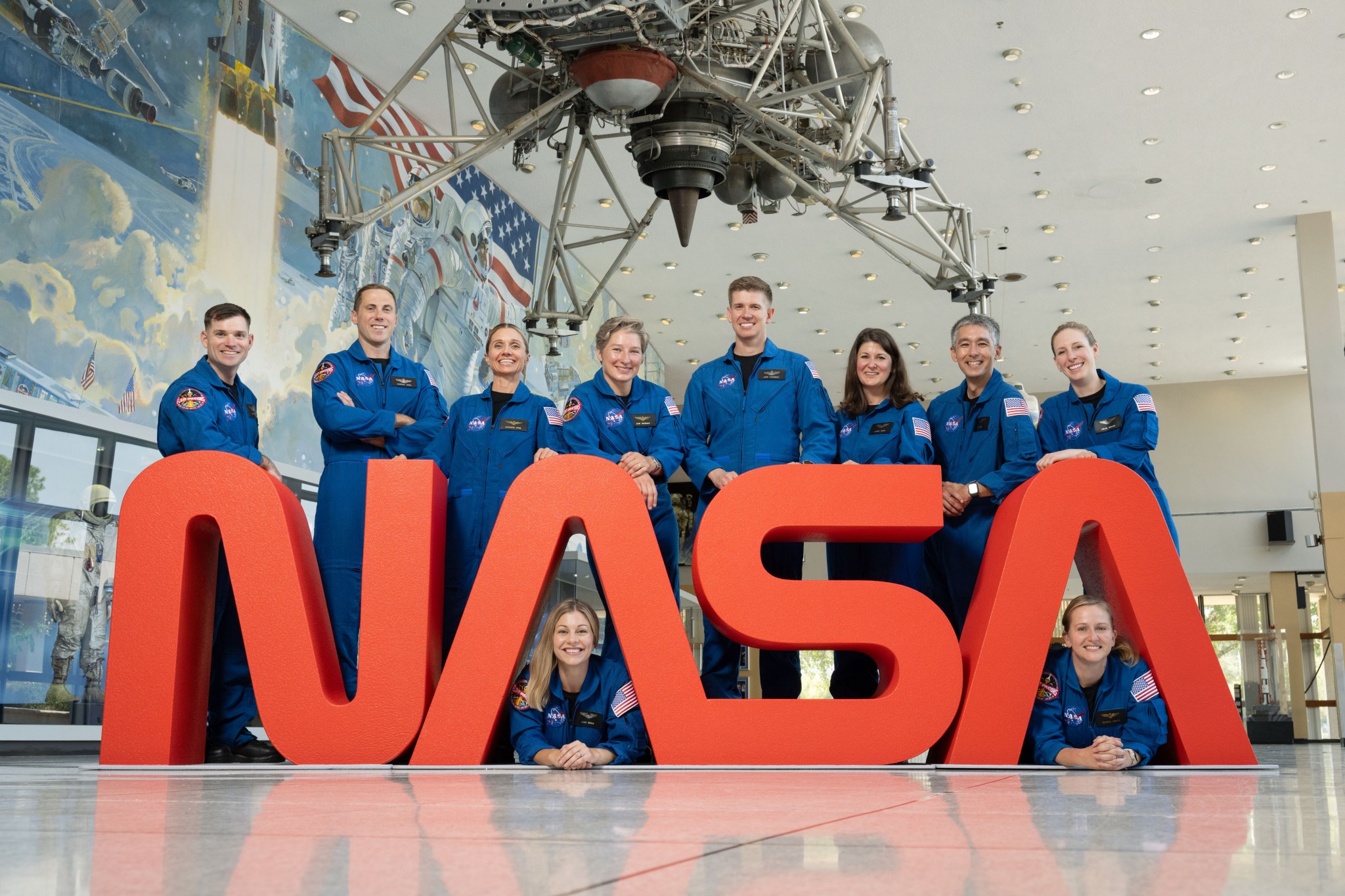 Ten members of NASA’s astronaut class in blue flight suits look and smile at the camera while gathered around a large NASA Worm logo.
