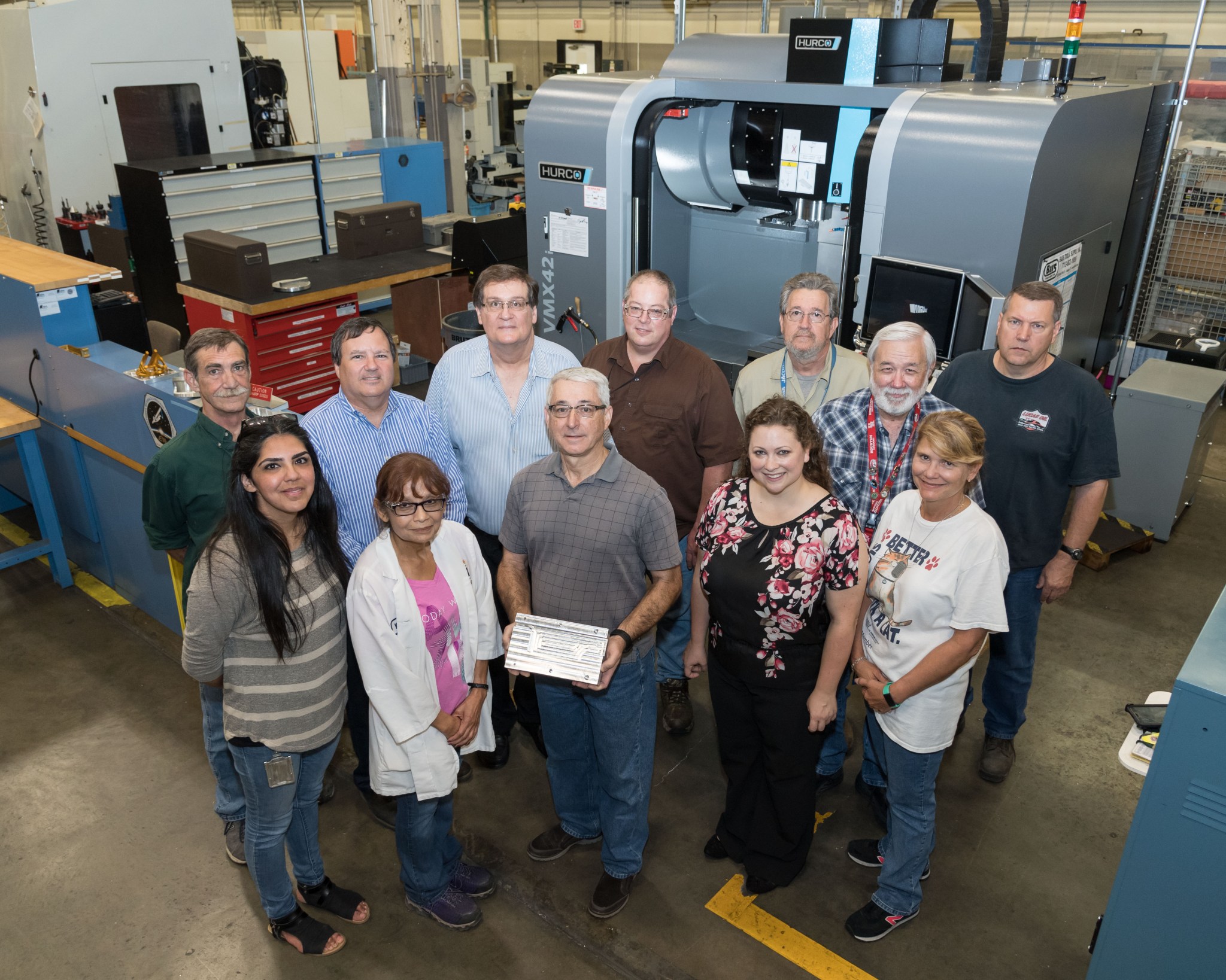 A group of twelve people pose in a facility with a miachie behind them.