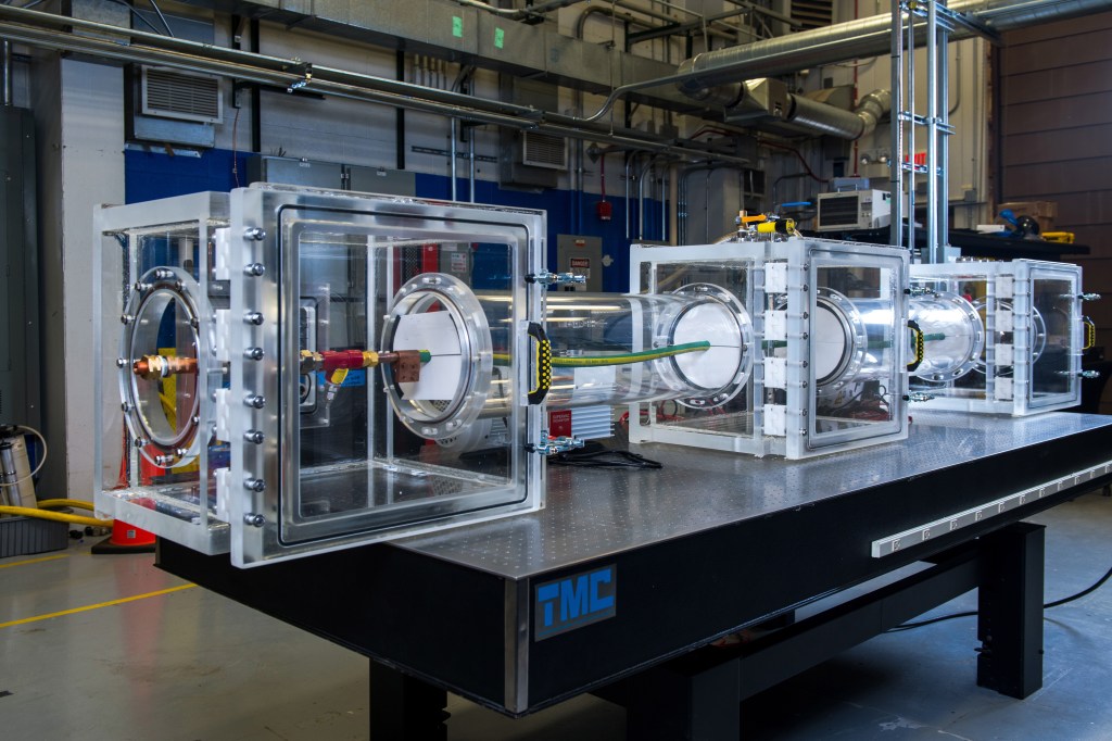 Three see-through boxes connected by a clear tube with wires and cables running through sit on top of a long table. The rig is surrounded by various hardware and machinery in the lab.