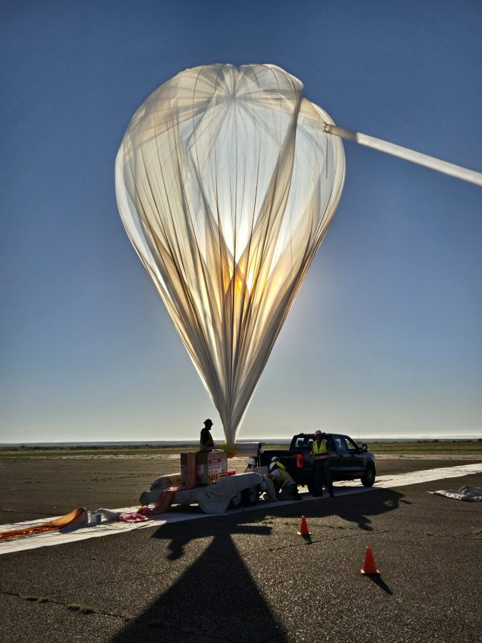 A large scientific balloon being inflated with helium during launch preparations, with ground crew and support vehicles visible below against a clear blue sky