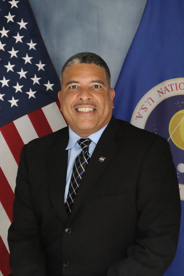 Image shows man in black suit posing for a photo with the American flag and NASA flag behind him