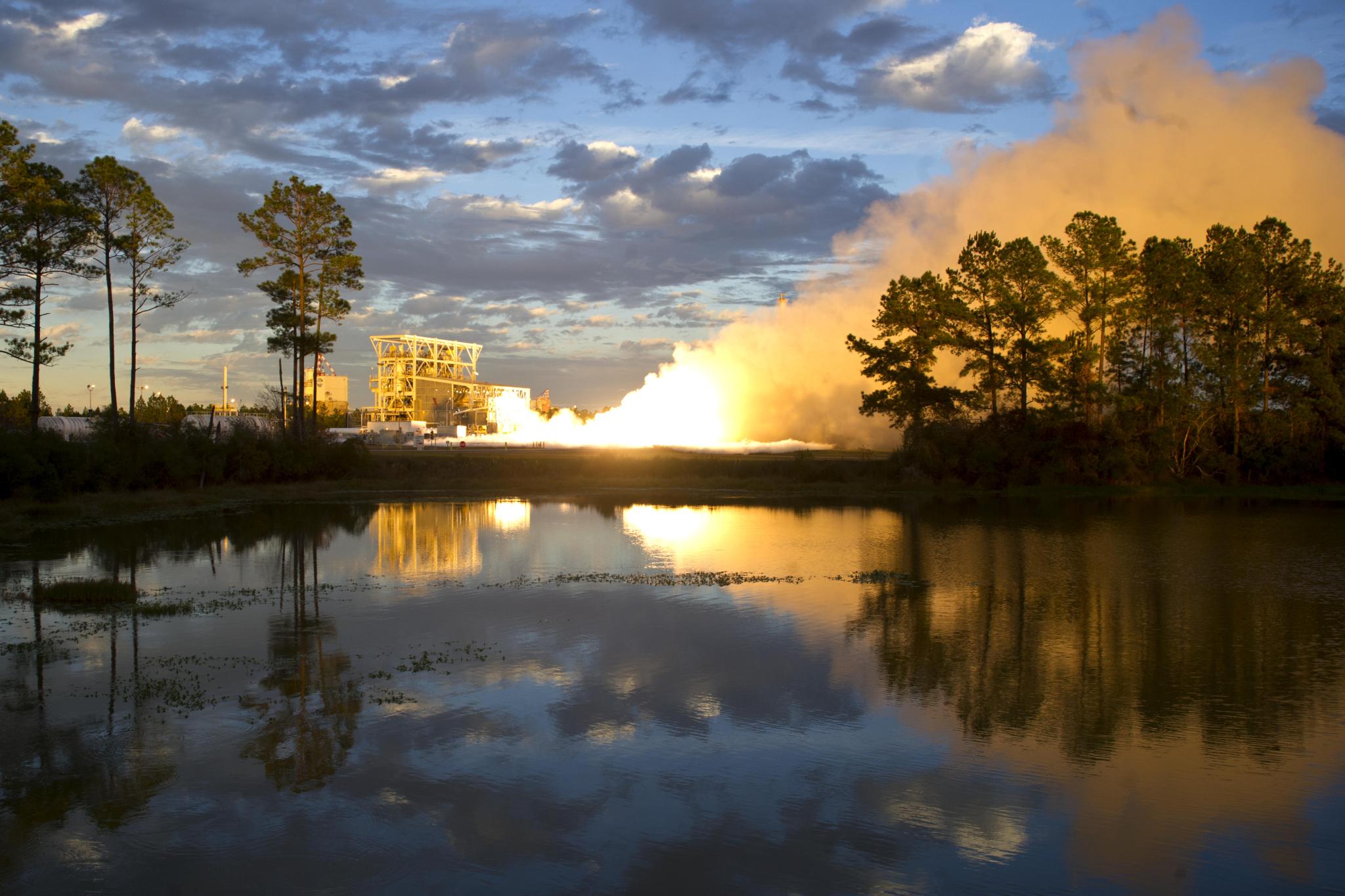 vapor clouds are aglow and reflected on a small body of water as the sun sets during test firing at Stennis Space Center