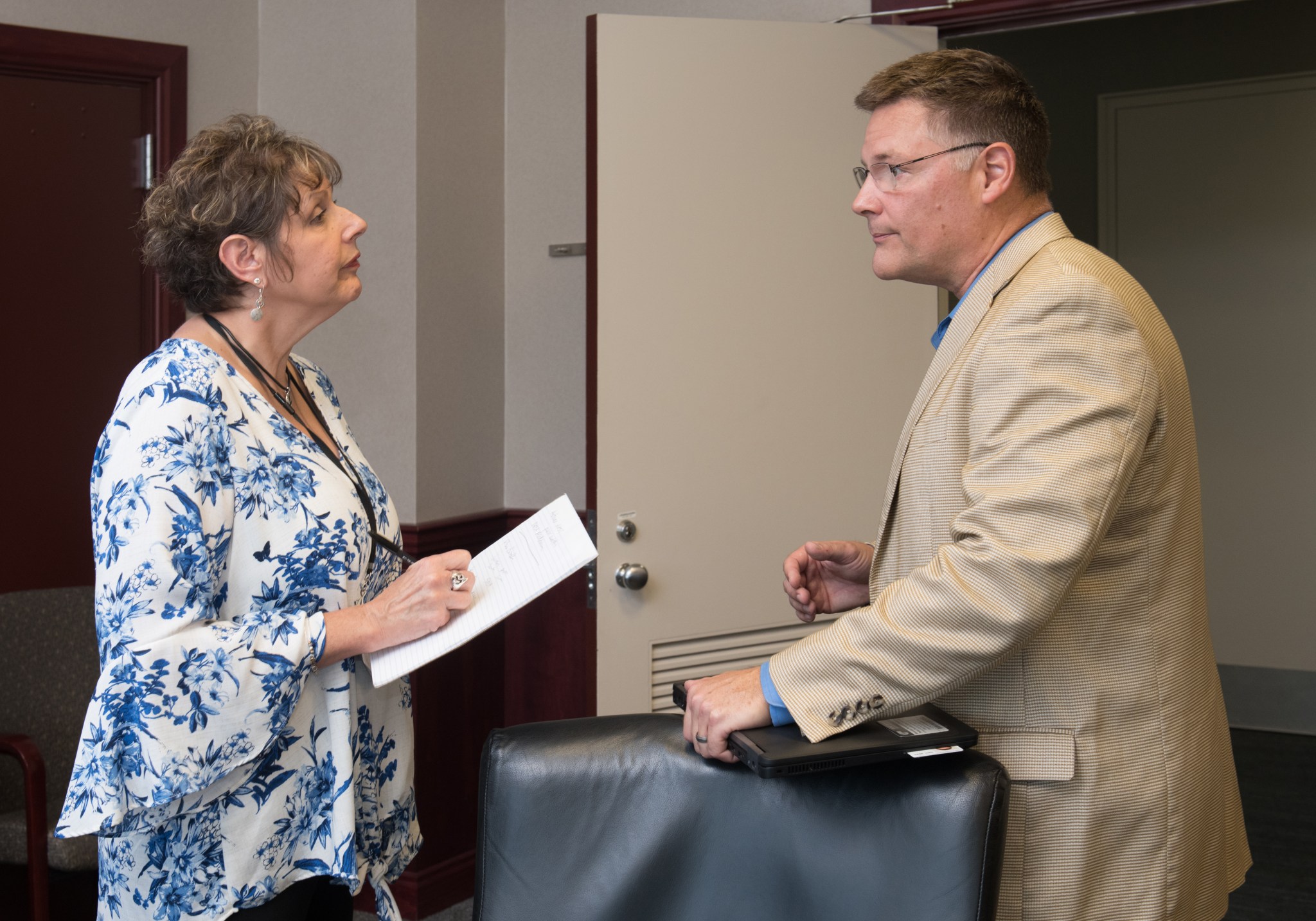 A woman takes notes on a large pad of paper as she faces a man wearing glasses and a tan suit. They stand in an indoor office space, and the man clutches a laptop in his left hand.