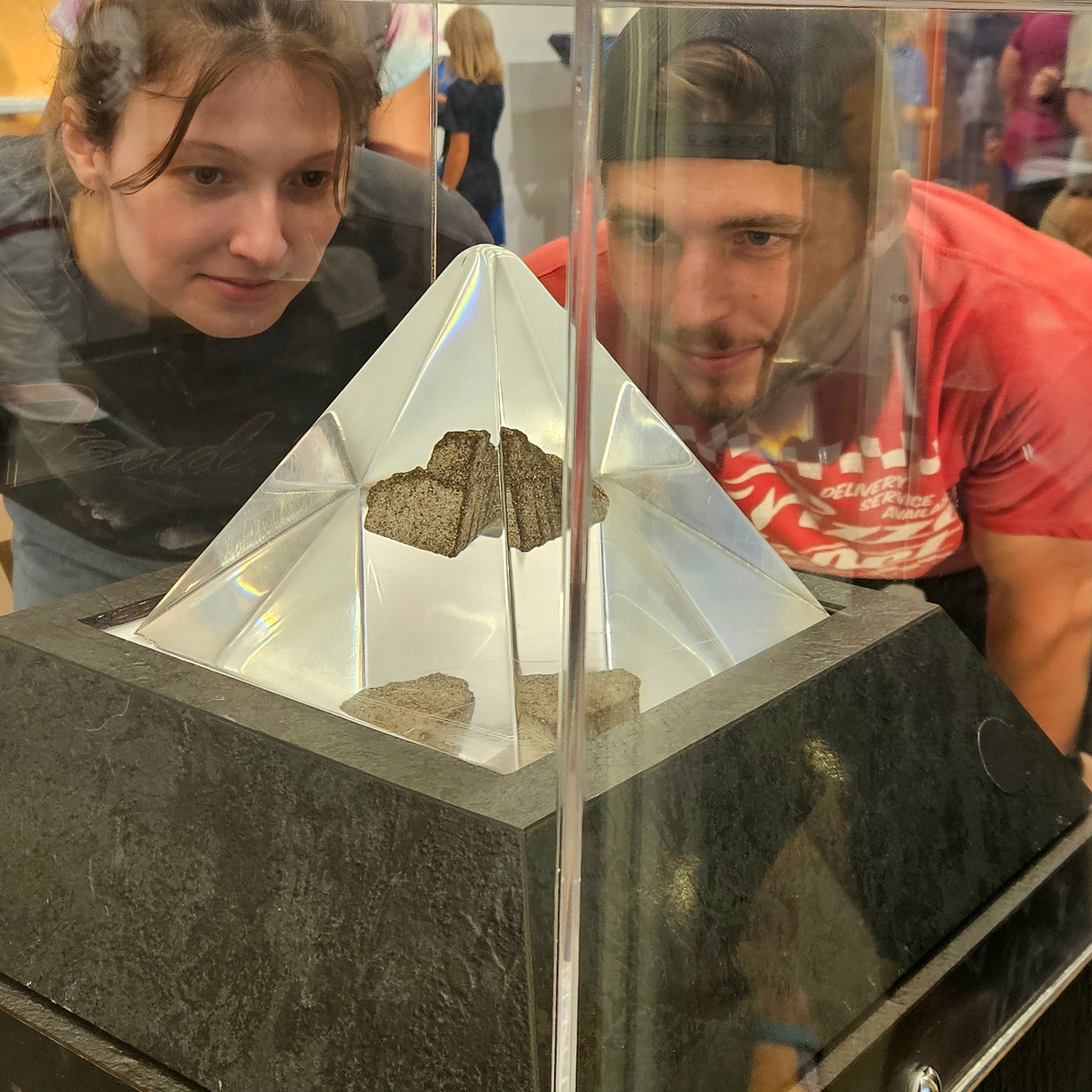 Two people crouch down to stare into an enclosed clear, polyethylene case containing a piece of a Moon rock.