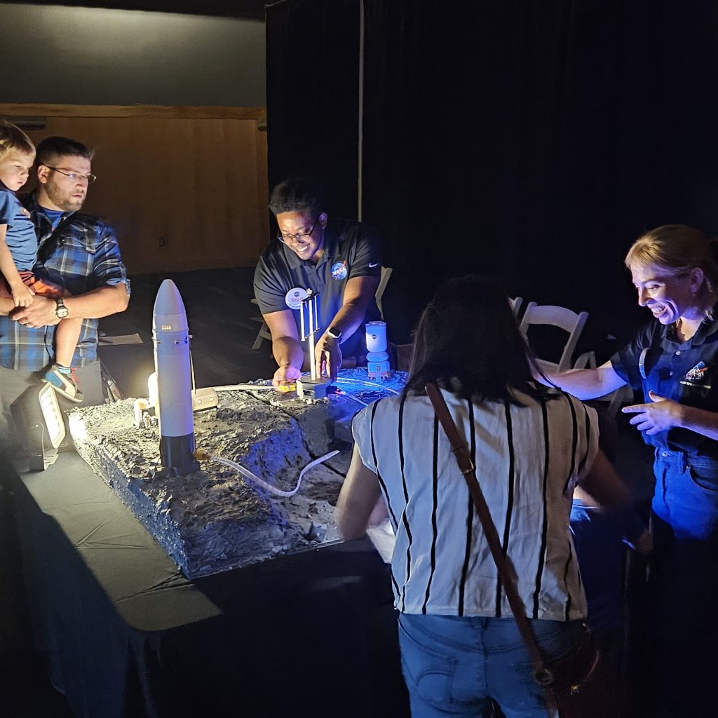 A NASA Glenn staff member connects lighted tubes to a tower on a grid that contains a rocket on a Moon-like surface. Guests and another staff member stand around the grid.