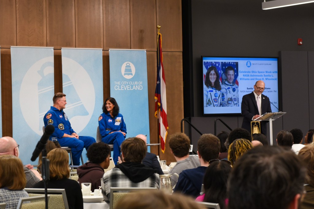 Two astronauts sit on stage in front of a backdrop that reads “The City Club of Cleveland.” To their right, the NASA Glenn center director stands at a podium and addresses the audience. Behind him is a screen with the astronauts’ photos and addresses the audience.