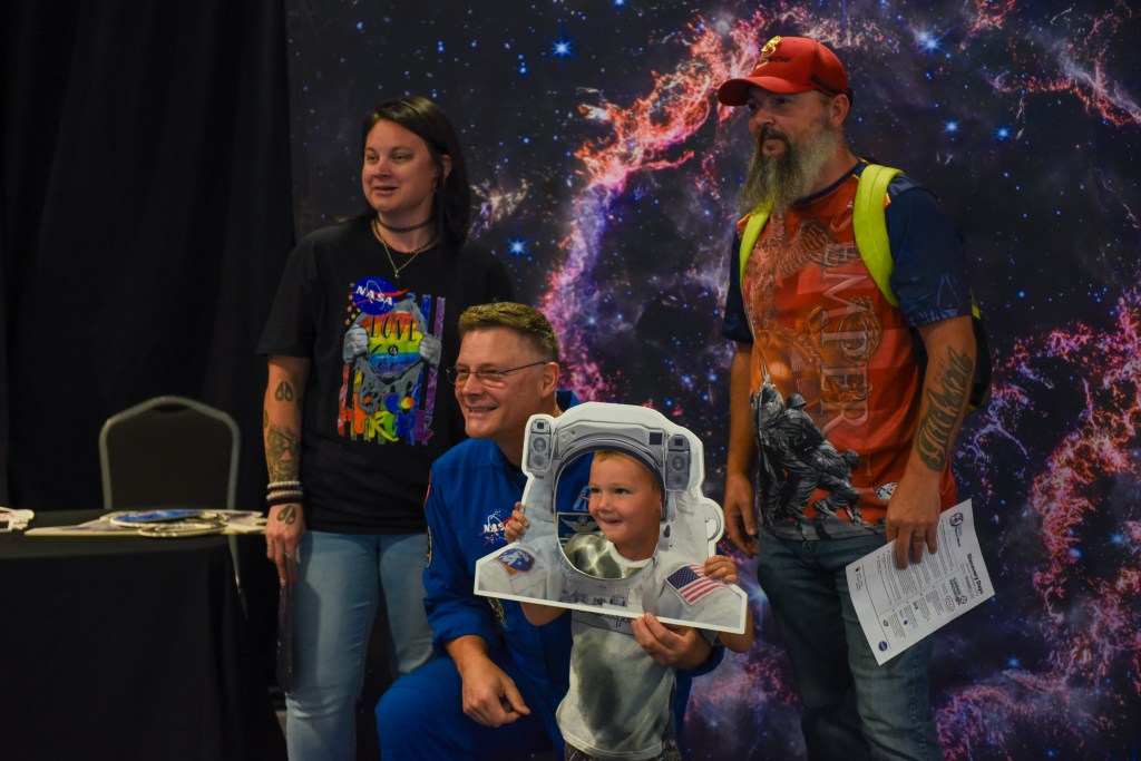 An astronaut crouches beside a young boy wearing a cut-out astronaut helmet. His parents stand beside them and in front of a cosmic backdrop and pose for a photograph.