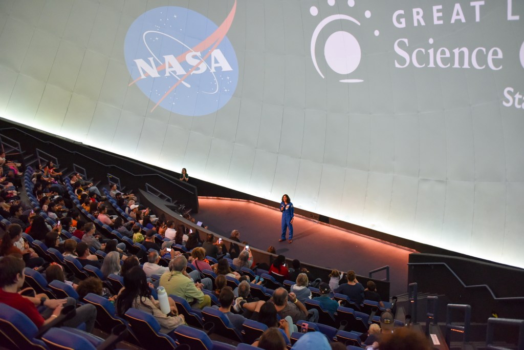 An astronaut stands at the bottom of a domed theater and waves at a crowd of seated students.