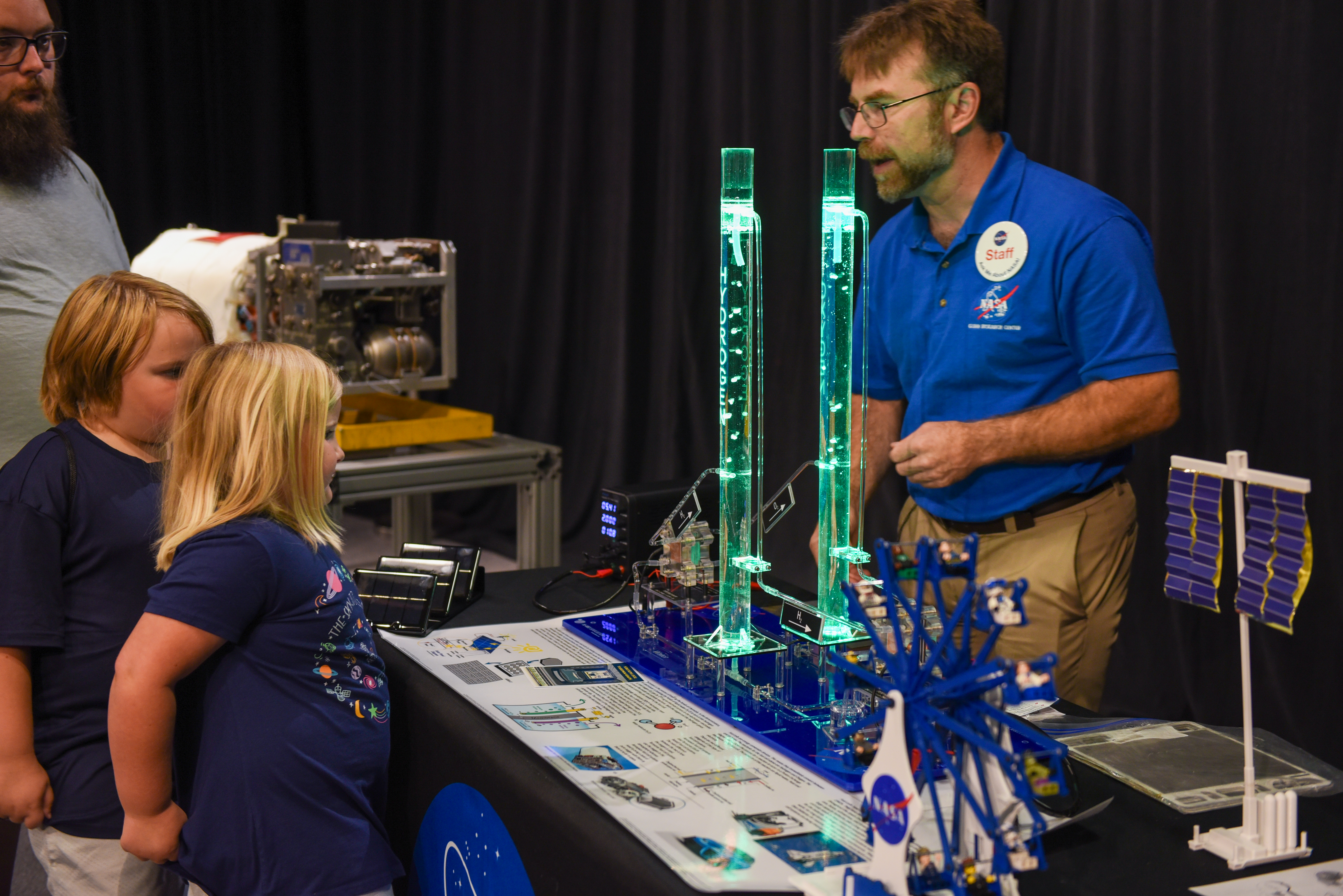 A NASA staff member stands behind a table containing two green tubes of liquid with bubbles while talking with two young visitors and their father.