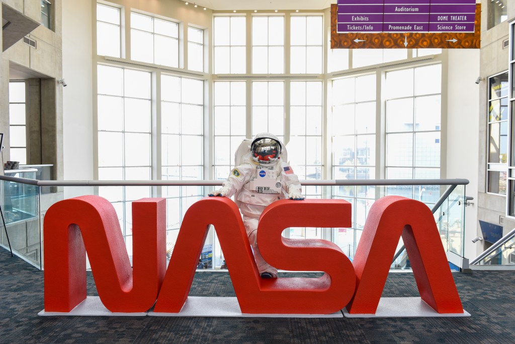 An astronaut mascot stands behind large red letters spelling out “NASA.”