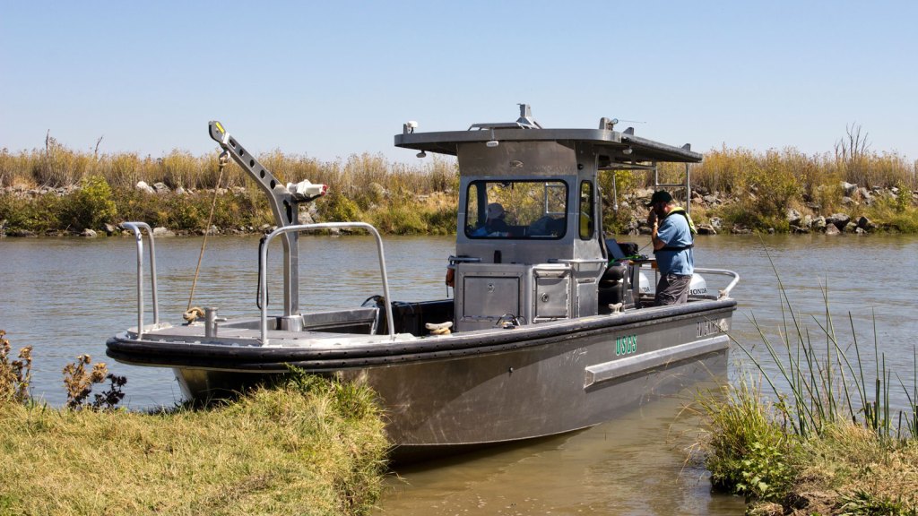 A silver-colored, flat-bottomed boat labeled "USGS" with a "Honda" outboard motor is docked at a grassy riverbank under a clear blue sky. A person in a blue shirt and yellow life vest is standing at the stern of the boat, looking at equipment. The boat has a small cabin and a crane arm at its bow. The river flows calmly, and both banks are lined with dry grasses and shrubs.