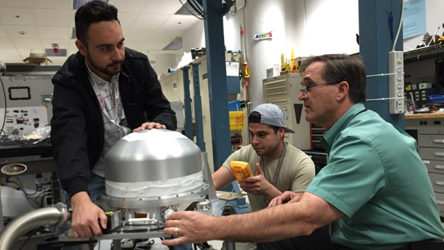 Three men are working on a piece of scientific equipment in a laboratory setting. In the foreground, a man with a beard and dark jacket is adjusting a large, silver, dome-shaped instrument. To his right, another man in a grey t-shirt and backward baseball cap holds a yellow multimeter and looks at the instrument. On the far right, an older man in a green short-sleeved collared shirt and glasses observes the work, engaged in discussion. The background shows various lab equipment and shelving.