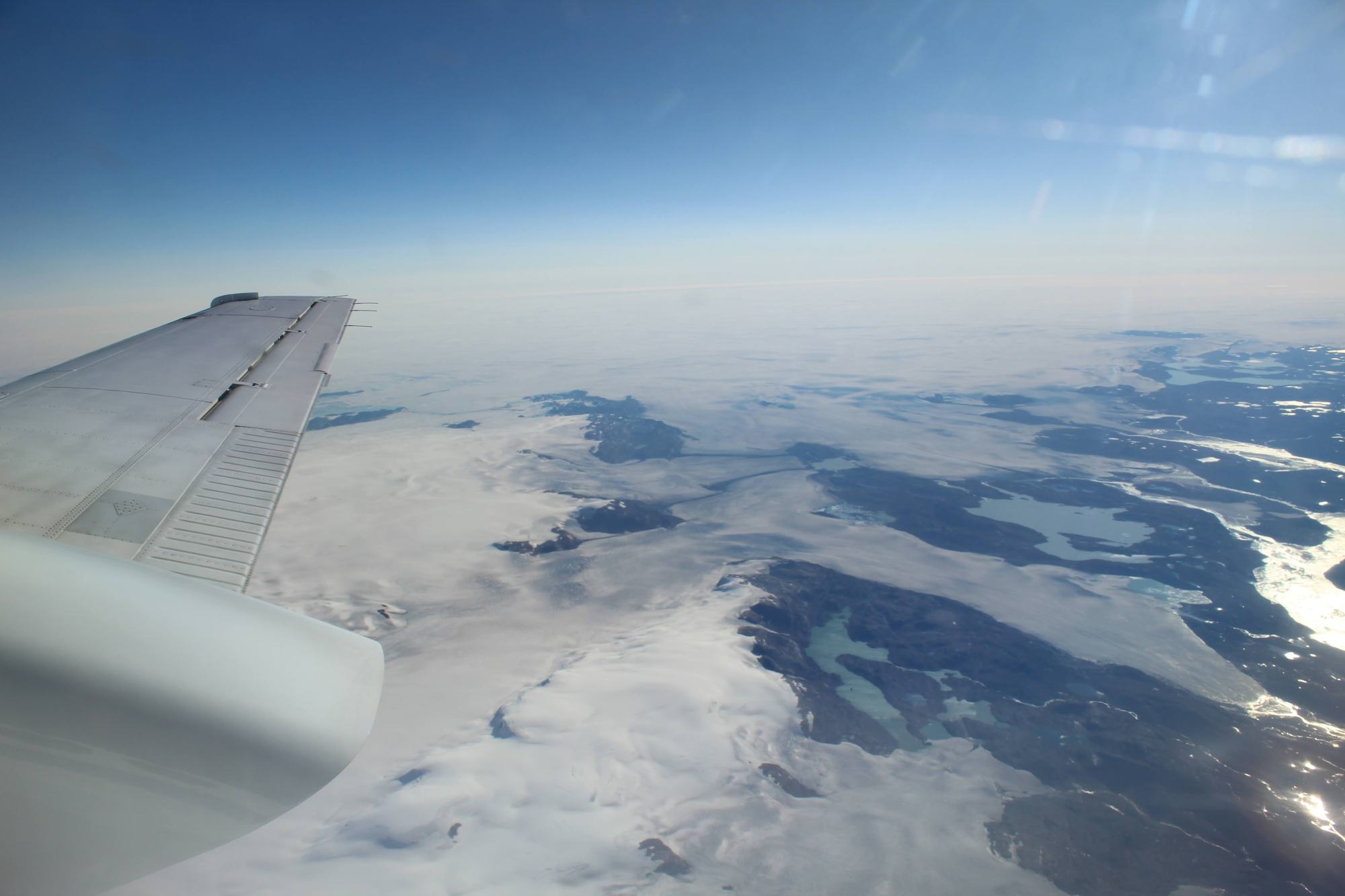 A view out of the window of the B200 King Air aircraft. A blue sky is seen at the top, then a cloud layer and water, ice and rock below. The tip of the plane is visible on the left.