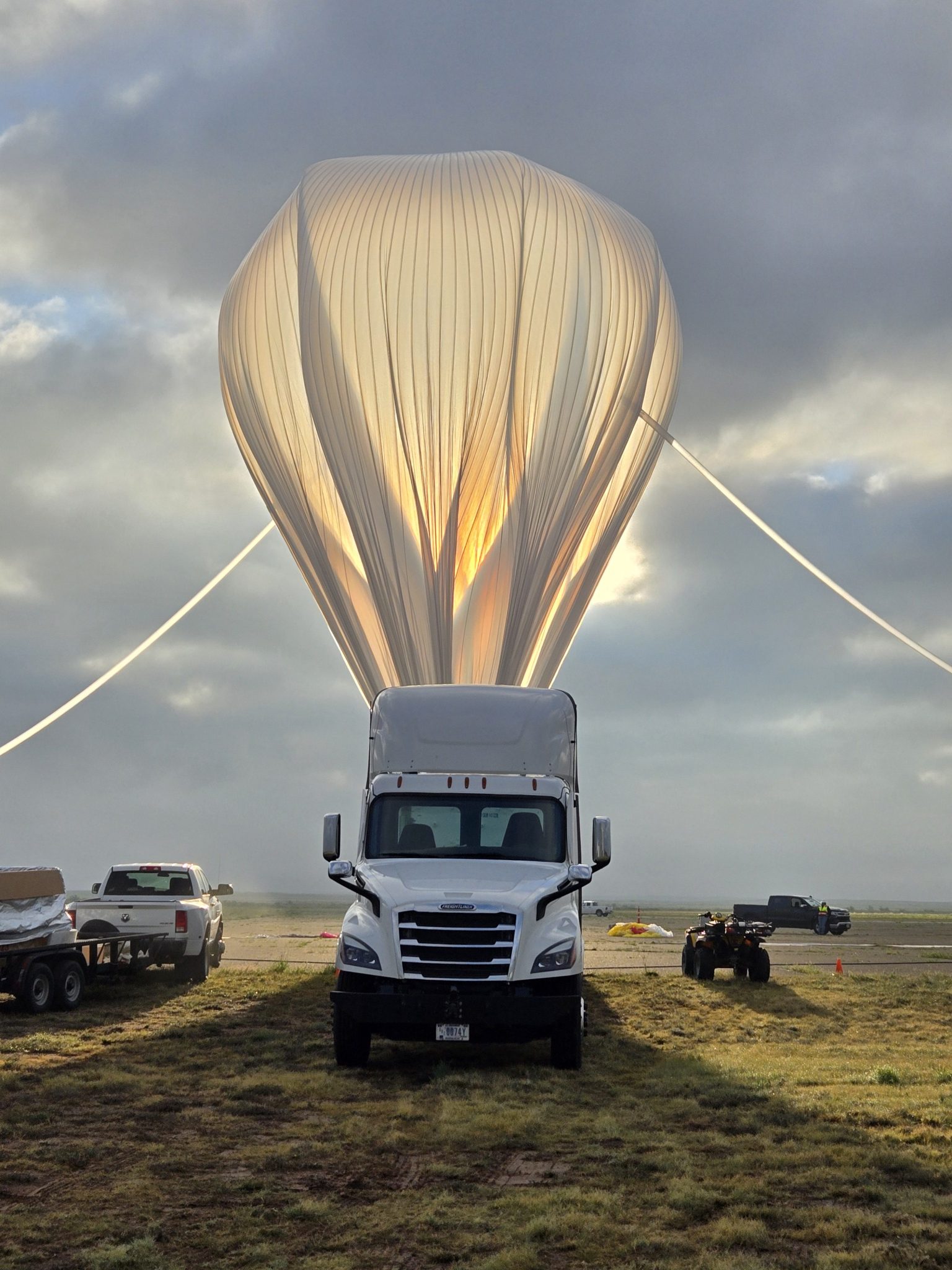 A NASA scientific balloon inflating behind a white truck during launch operations, with the balloon glowing in the sunlight against a cloudy sky.