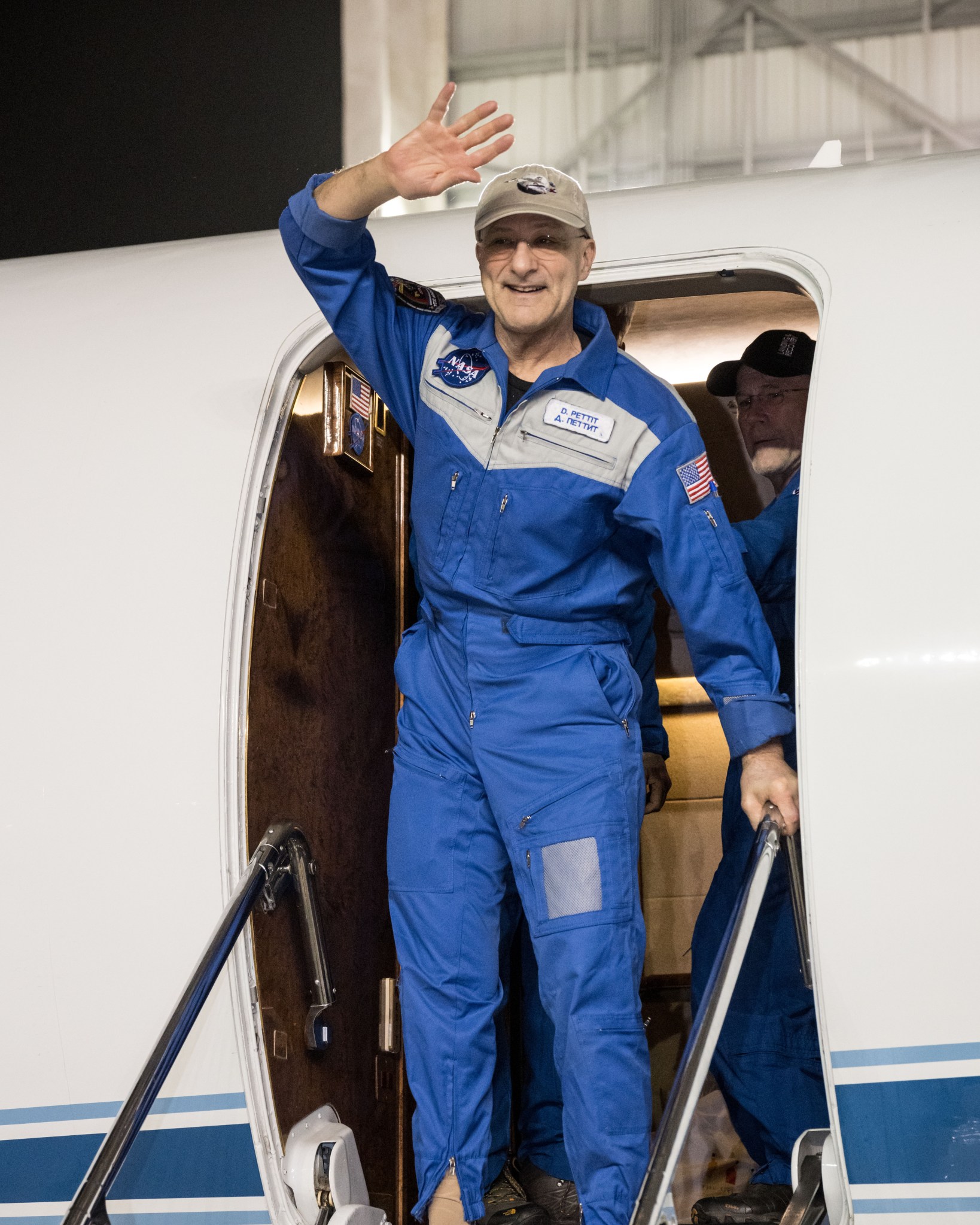 A man in a blue flight suit stands and waves as he exits a plane.