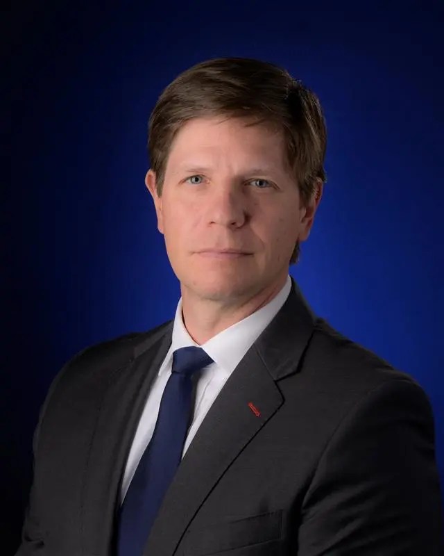 Portrait of Kevin Murphy, male, in front of a blue background wearing a black suit jacket and dark blue tie.