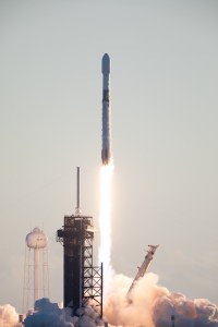 A rocket lifts off into the sky, leaving a bright trail of flame in its wake. White vapor clouds billow out along the ground. There are also various structures in the image, including a gantry and a white structure that looks like a water tower.