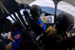 NASA astronauts Matthew Dominick (left, in a blue jumpsuit with a patch on the right arm) and Mark Vande Hei (right, in camouflage clothing) in the cockpit of a helicopter. Dominick holds an open binder in his right hand and points forward with his left. They are both wearing black helmets. There is another person behind Dominick, also wearing a blue jumpsuit and helmet, facing the opposite way.