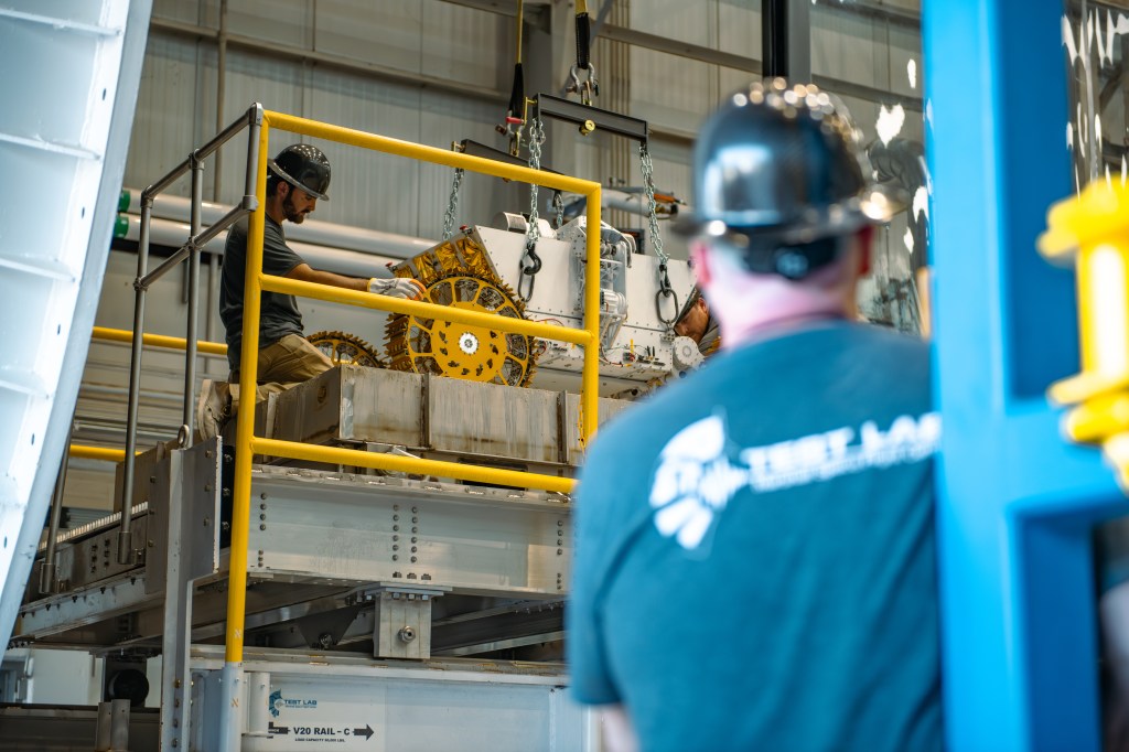 A technician wearing a black hard hat and gloves kneels on an elevated metal platform, carefully positioning a large robotic lunar rover into place. The rover has gold-colored wheels with a complex tread design and is suspended by heavy-duty chains and straps attached to a lifting rig. Another individual in the foreground watches the procedure closely. The scene takes place inside an industrial testing facility with metal scaffolding, safety rails, and visible structural supports.