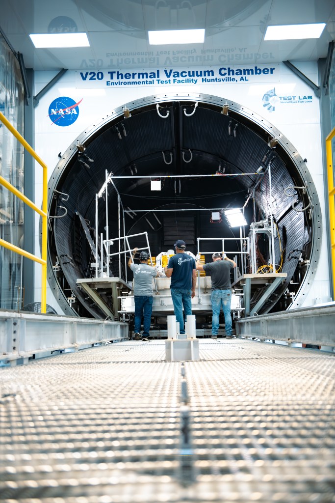 Three people wearing hard hats stand on a grated metal walkway facing a large circular vacuum chamber with its door open. They are working near a metal platform inside the chamber. The scene includes yellow guardrails sidelining the walkway, overhead lights reflecting on white panels, and cables and metal structures inside the chamber. The vacuum chamber’s door features the NASA “meatball” insignia next to the words “V20 Thermal Vacuum Chamber Environmental Test Facility Huntsville, AL.”