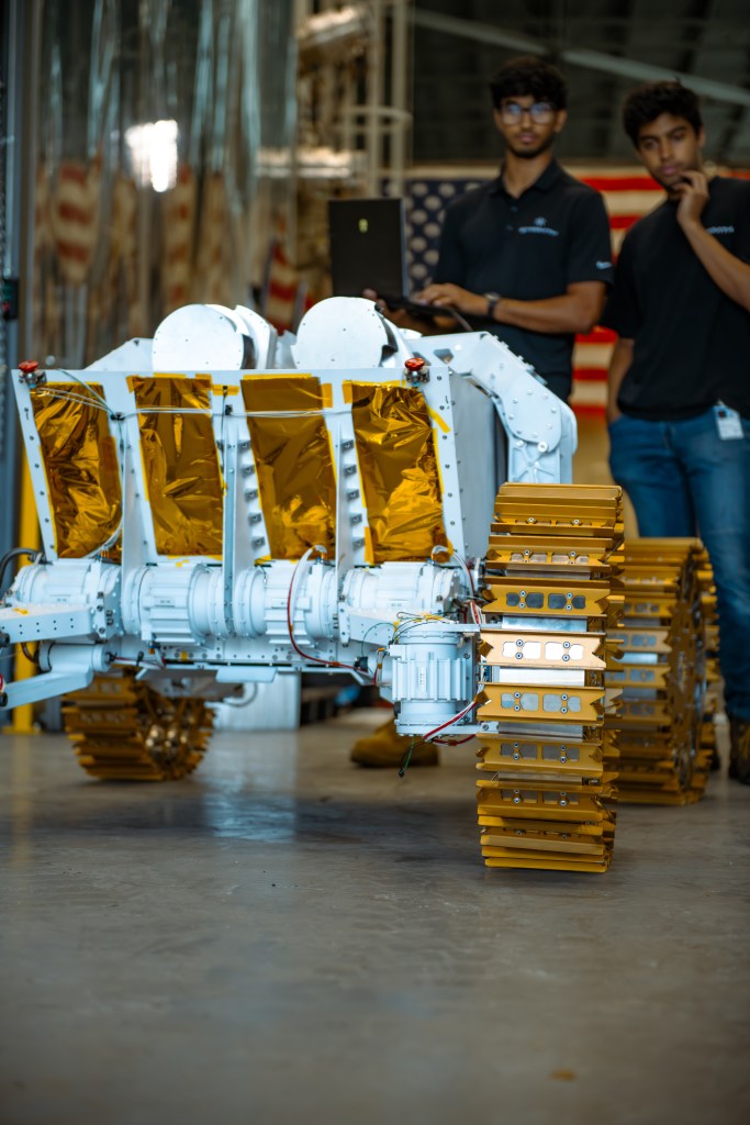 A close-up view of a robotic lunar rover with golden metal treads and a white body, featuring exposed wiring and gold thermal insulation panels. The rover is indoors on a smooth concrete floor. Behind the rover, two individuals wearing dark polo shirts and jeans are standing, one holding a laptop and the other with a hand near his face. An American flag is partially visible in the background. The focus is on the rover, with the people and background slightly out of focus.