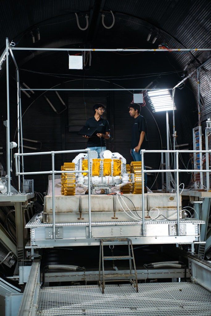 Two people wearing dark shirts stand on a metal platform inside a large vacuum chamber, with bright lights mounted nearby. They are next to a large lunar rover, featuring gold-colored metal treads and white body panels with reflective gold thermal insulation. The platform has metal railings, cables running along its base, and a step ladder leading down to a grated walkway.