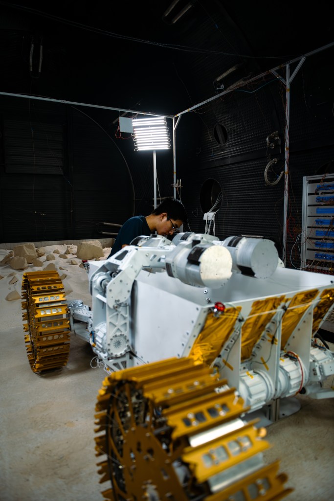 A person works on a rugged robotic vehicle inside a dimly lit chamber. The robot features gold metallic wheels with deep tread patterns, exposed gears, and motors, and sits atop a concrete slab. The workspace is cluttered with cables and equipment, and a bright overhead light illuminates the area. Sand and rocks scattered on the floor indicate a testing environment simulating planetary surfaces.