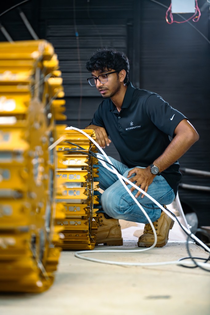 A person kneels beside a large, metallic rover with a golden finish, inspecting it. The robot, whose large golden wheels can be seen just out of focus, is connected to multiple cables as it prepares for testing. The individual wears a black polo shirt, jeans, and tan work boots. The setting is a thermal vacuum chamber with a dark wall and visible wiring, and a concrete floor.
