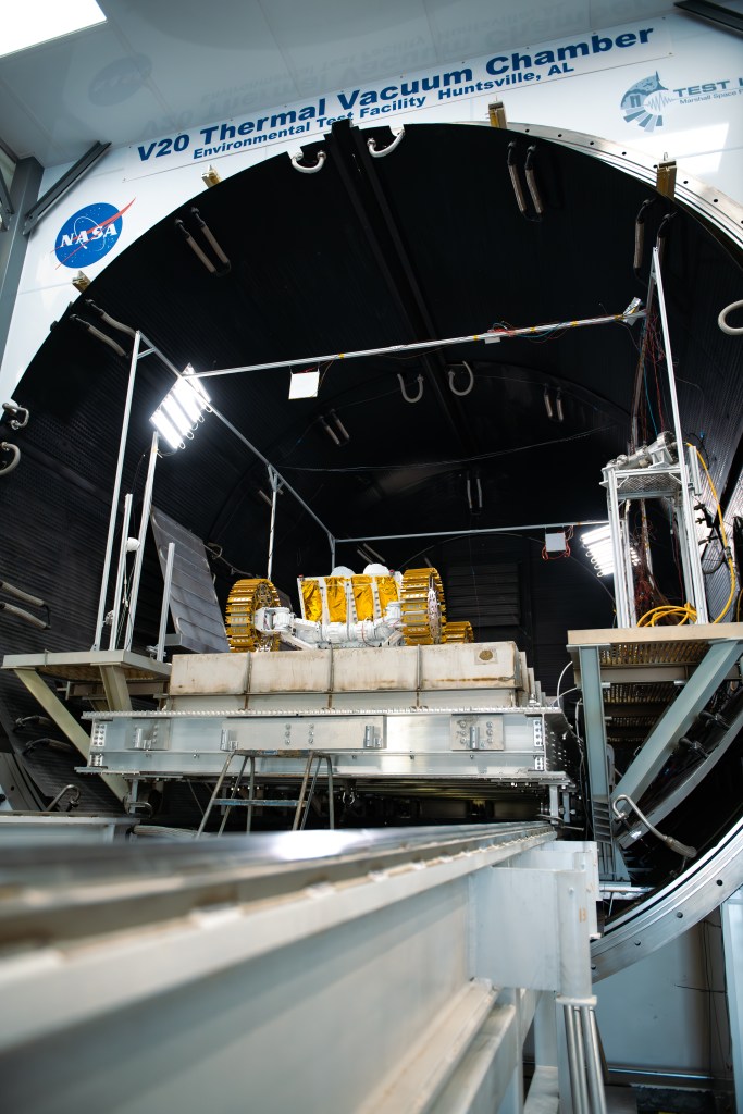 A large cylindrical chamber labeled "V20 Thermal Vacuum Chamber" with the NASA “meatball” insignia next to it is shown in an indoor testing facility. Inside the chamber, a robotic lunar rover prototype can be seen sitting atop a concrete slab on a sliding platform. The robot features golden metal treads on its four large wheels and a white body, surrounded by cables and structural supports.