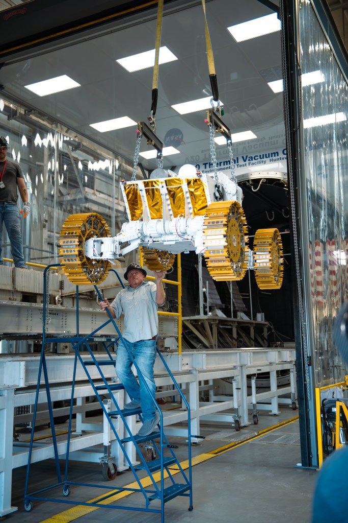 A white and gold lunar rover prototype is suspended mid-air by heavy-duty straps and chains attached to a ceiling-mounted crane system inside a clean, industrial facility. The rover, featuring gold-colored metal treads and white body panels with reflective gold thermal insulation, is being carefully guided by two workers. One man stands on a rolling blue metal staircase, using a pole to stabilize the rover. Another worker observes nearby on an elevated platform. The background includes a large thermal vacuum chamber. The setting is well-lit with overhead fluorescent lights, and parts of an American flag are visible through transparent plastic curtains framing the entrance of the thermal vacuum chamber.