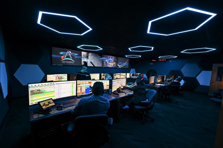 A dark room with 8 scientists in front of computers with multiple monitors. At the front of the room, there are 4 large screens with the Artemis logo and mission data.