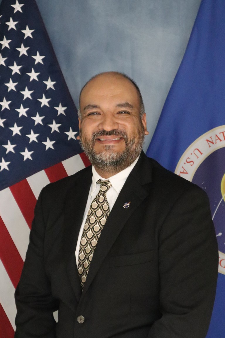 Image shows gentleman in a black suit posing for a photo with an American and NASA flags in the background