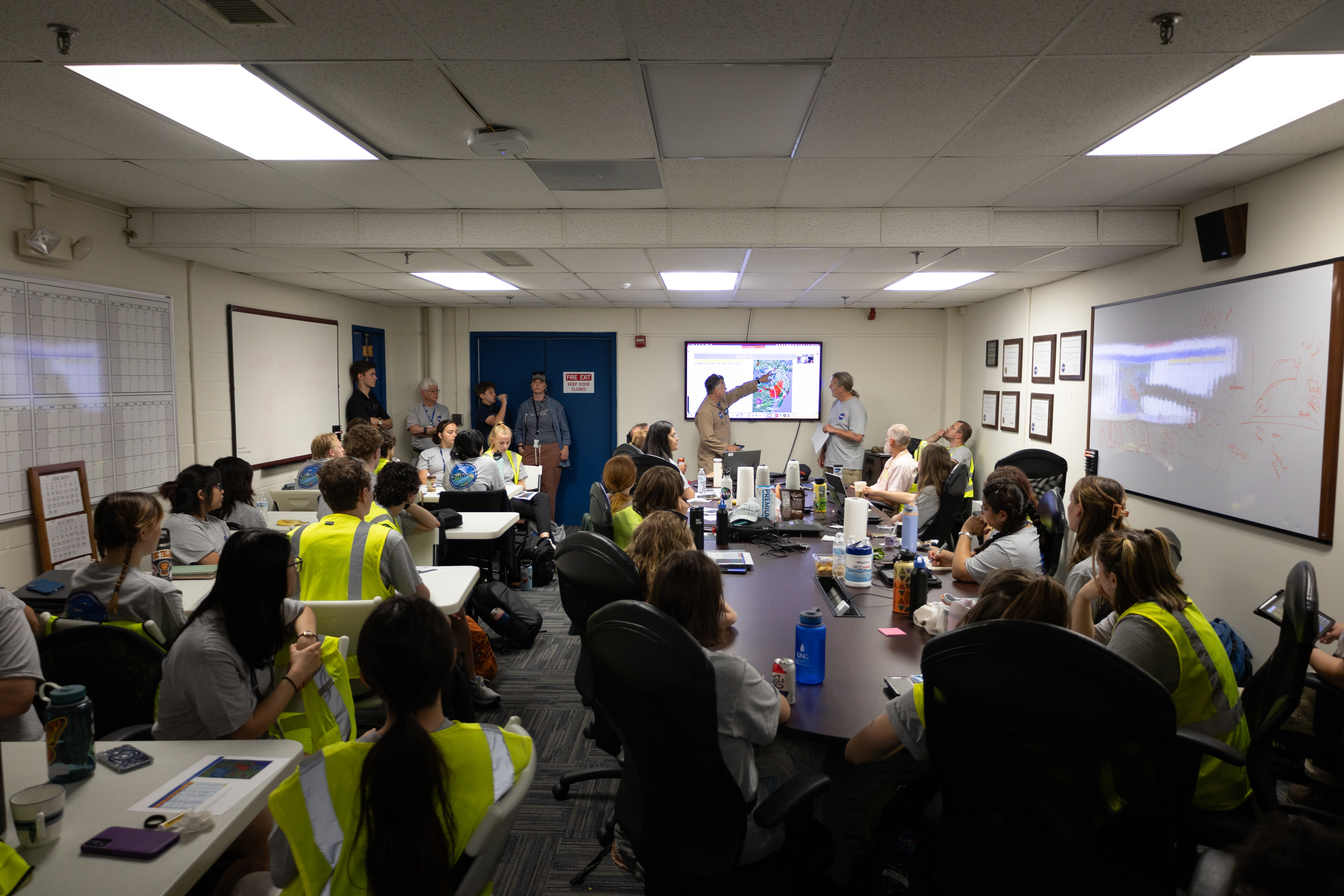 A man in a tan flight suit points at a screen with a graphic of the United States. In the foreground, a group of people sits around white tables, listening with their backs to the camera.