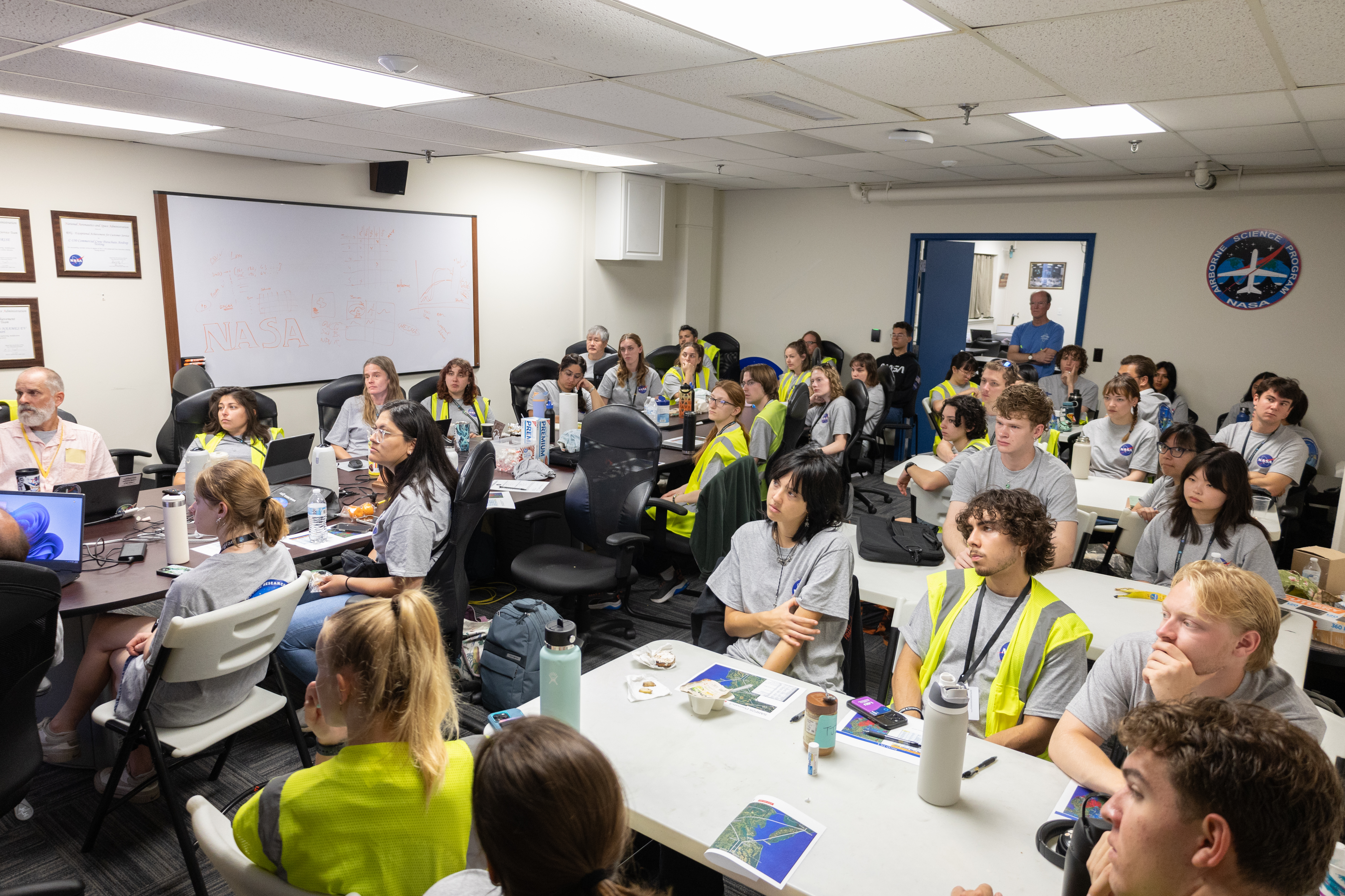 A classroom full of students and staff packed around white tables listen to someone out of the frame.