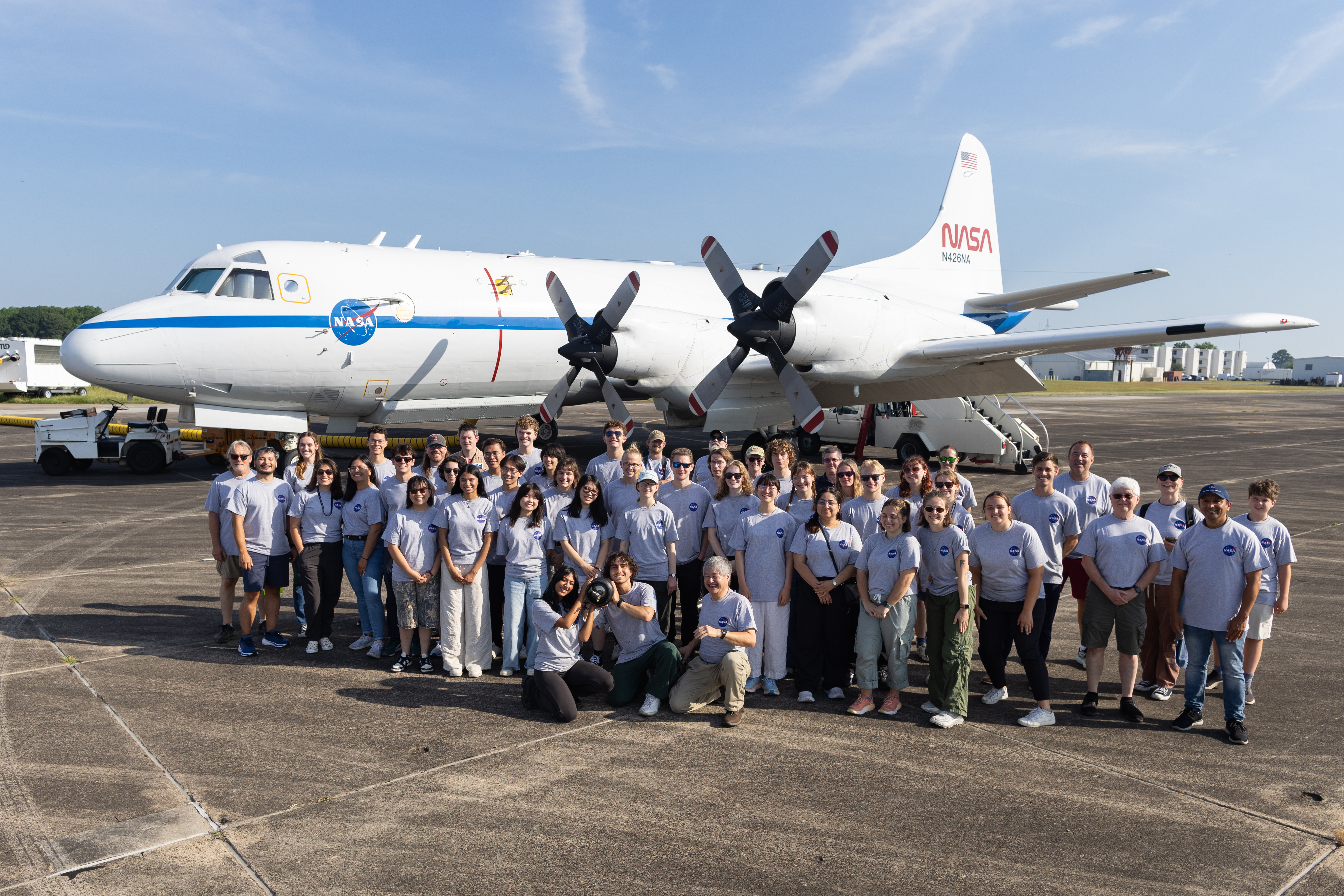 A group of students and faculty in gray shirts stand on dark gray tarmac in front of a large white plane with a blue stripe down the middle.