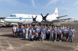 A group of students and faculty in gray shirts stand on dark gray tarmac in front of a large white plane with a blue stripe down the middle.