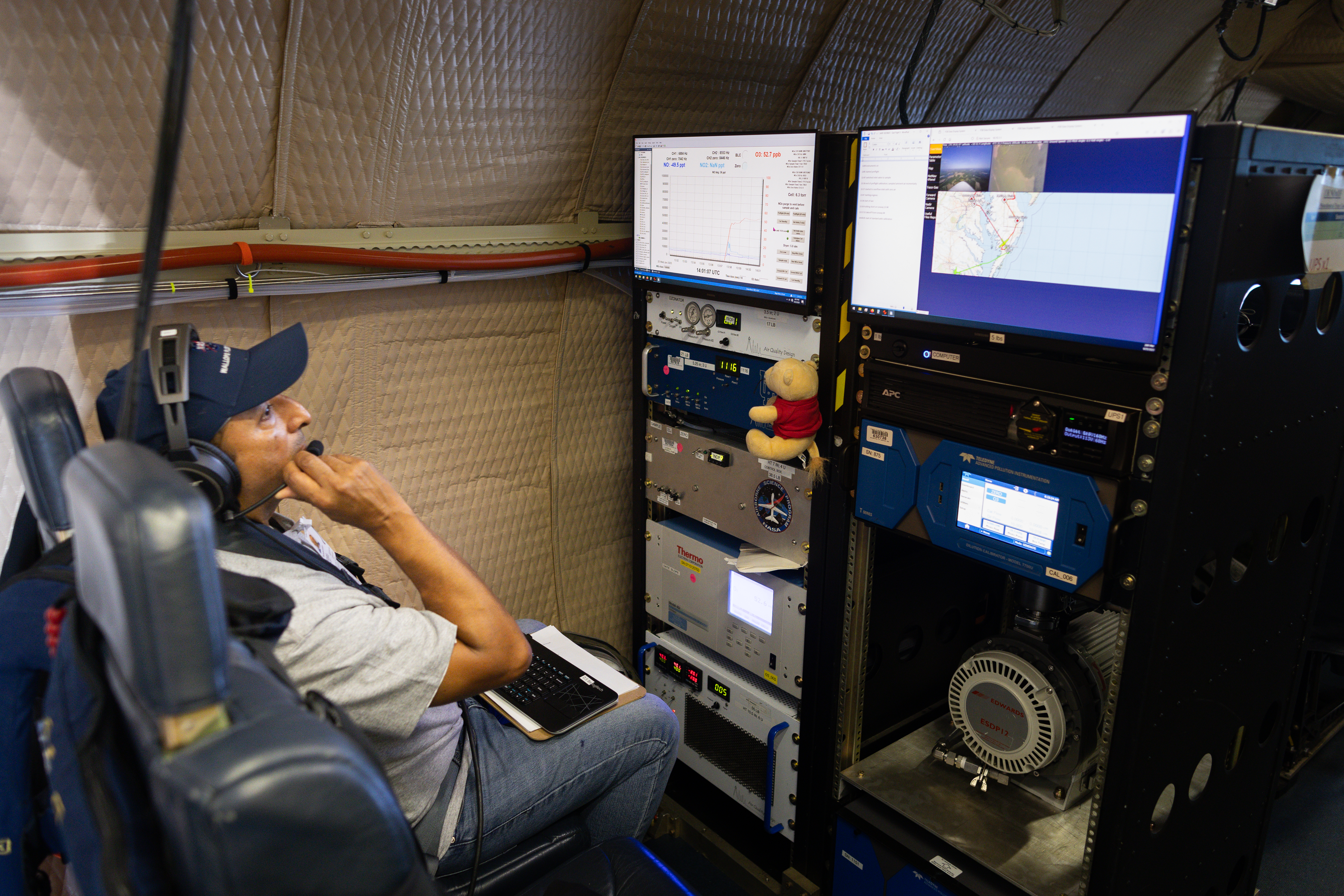 A man in a navy baseball cap, gray t-shirt, and headphones sits in a dark blue chair, looking at two screens.