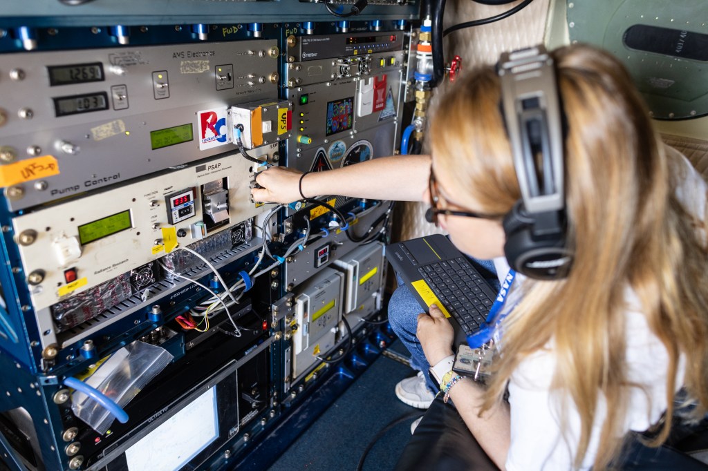 A woman with blonde hair, headphones, and glasses leans left to adjust dials on a large instrument in front of her.