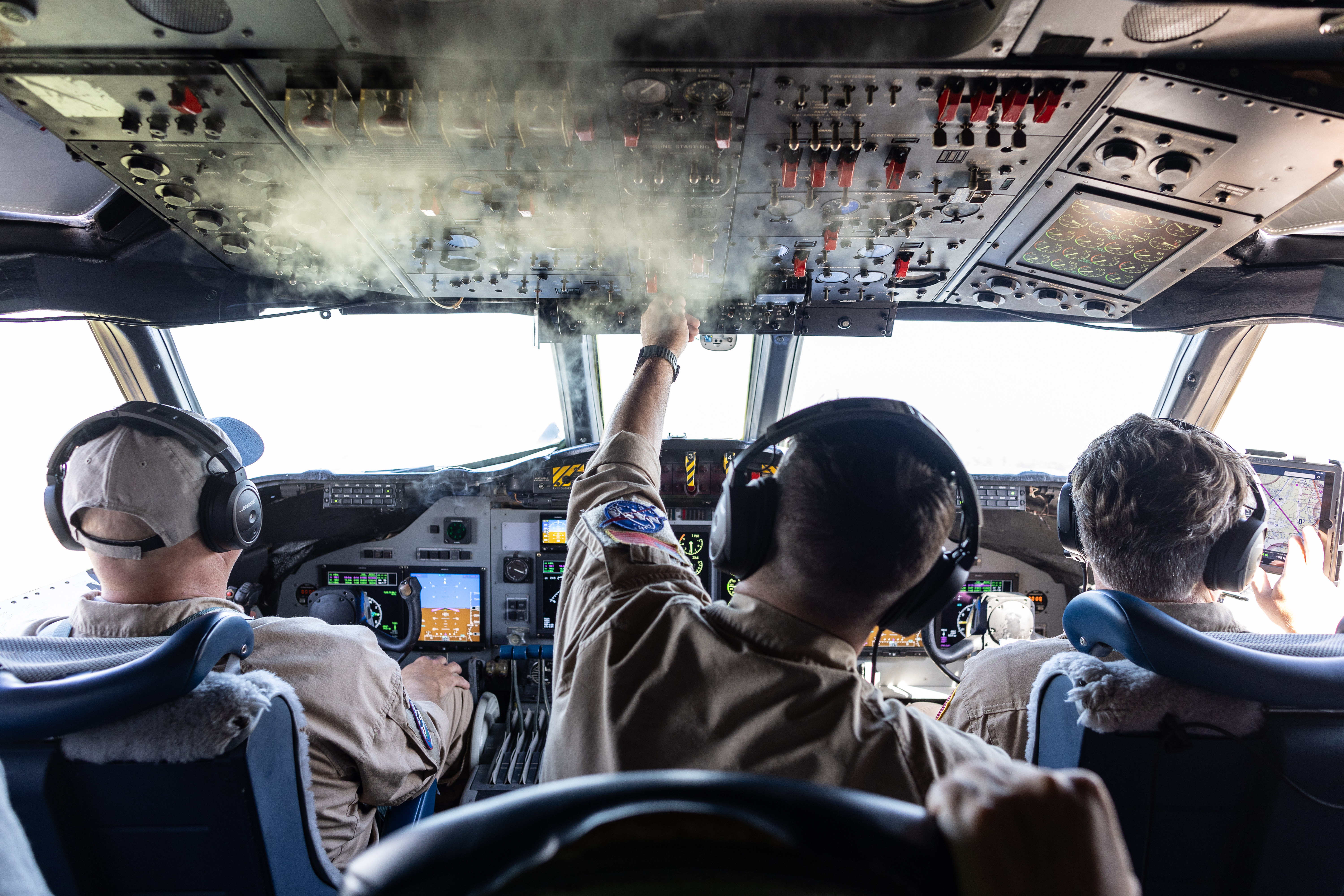 Three men in headphones sit in a cockpit with their backs to the camera, watching the displays. A bit of mist clings to the ceiling.