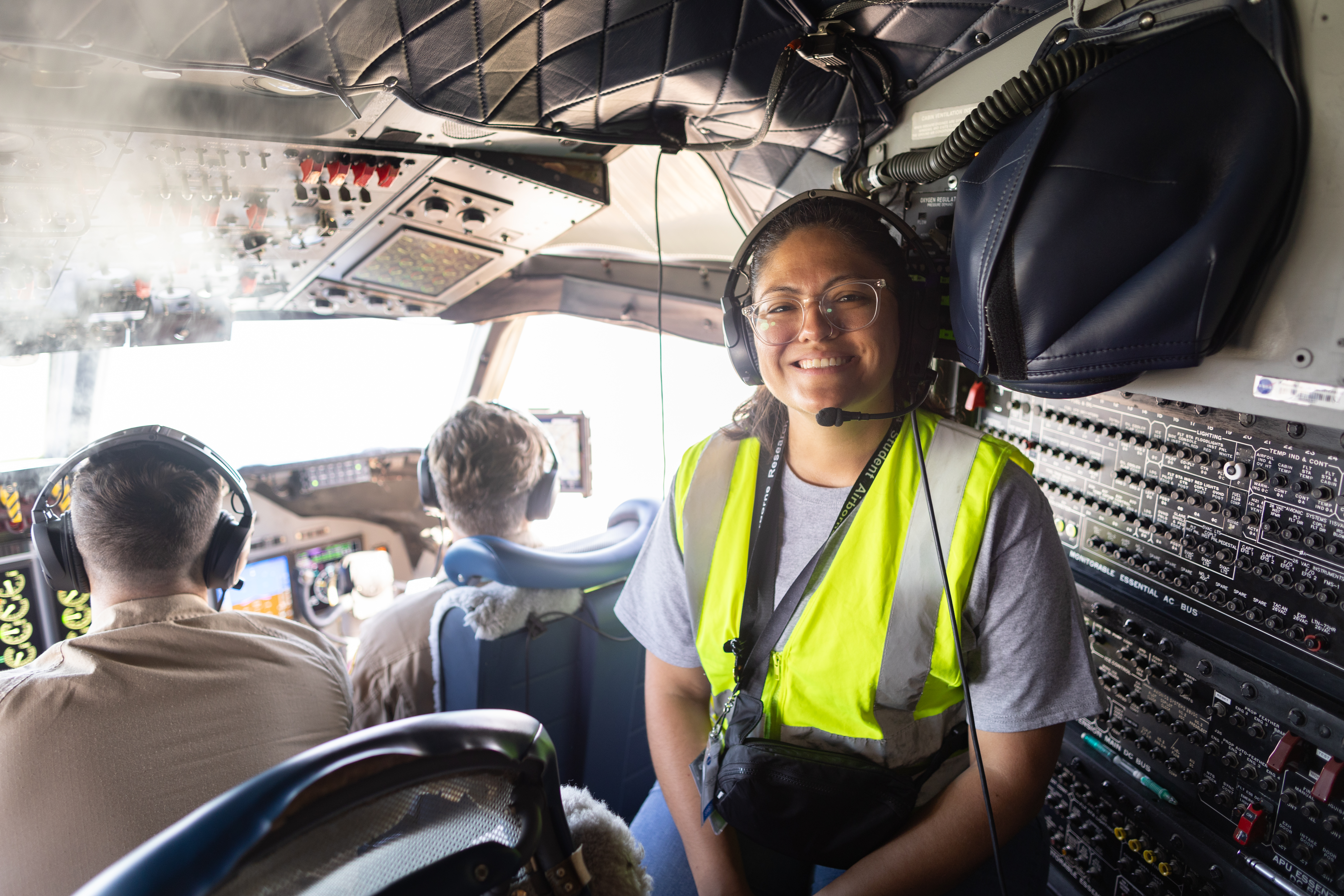A woman in a yellow high-viz vest and headphones smiles at the camera, sitting in the cockpit of an airplane.