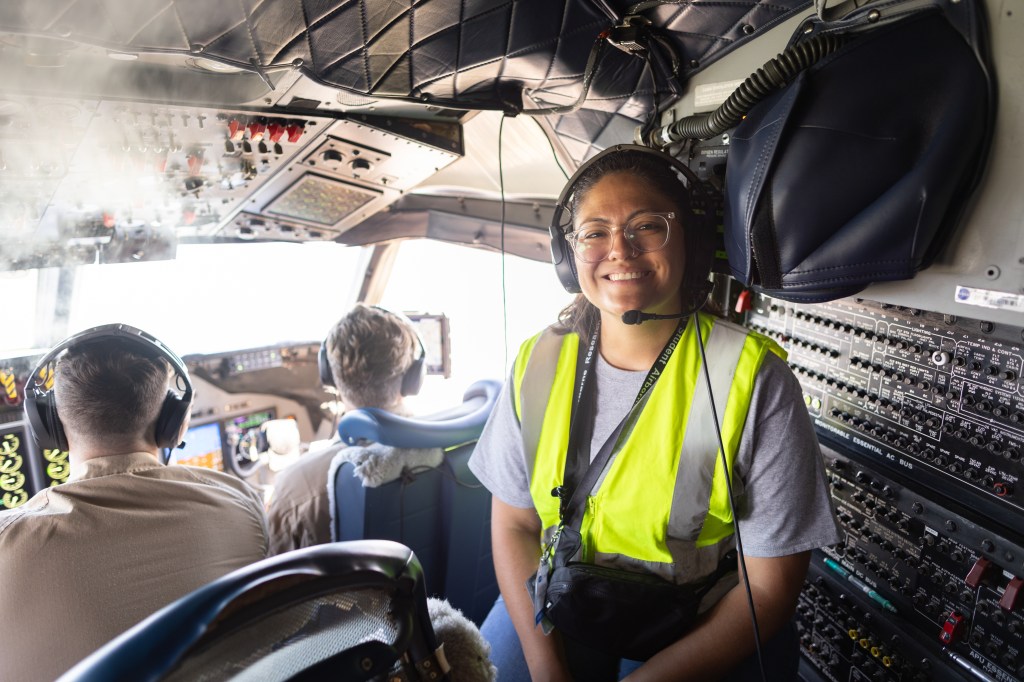 A woman in a yellow high-viz vest and headphones smiles at the camera, sitting in the cockpit of an airplane.