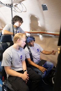 Two men in gray shirts and headphones sit looking at a screen. A student dressed the same stand behind them, leaning on the back of the chairs.
