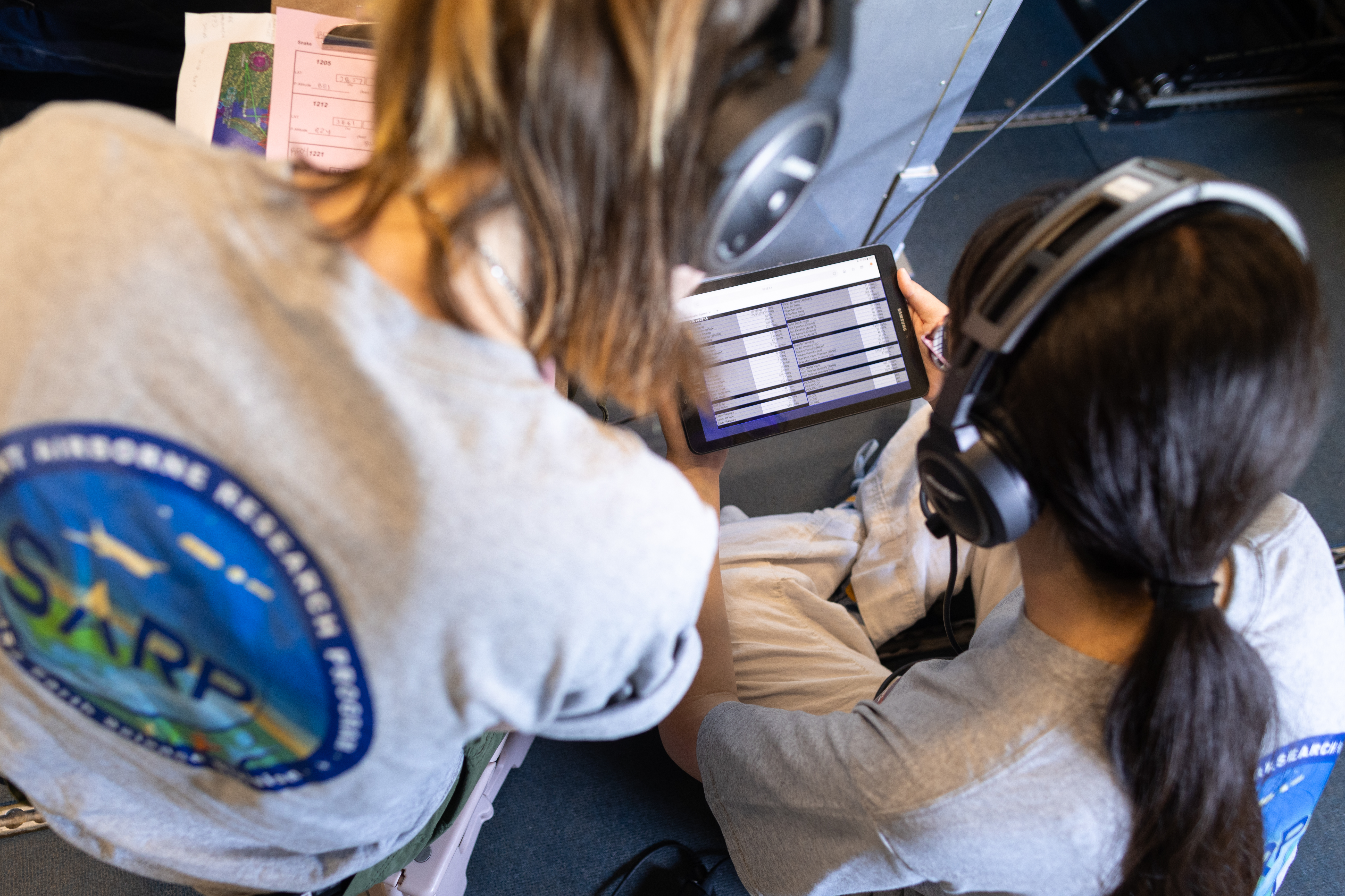 Two women wearing headphones and gray shirts with the SARP logo sit on the ground, looking at a tablet with data.