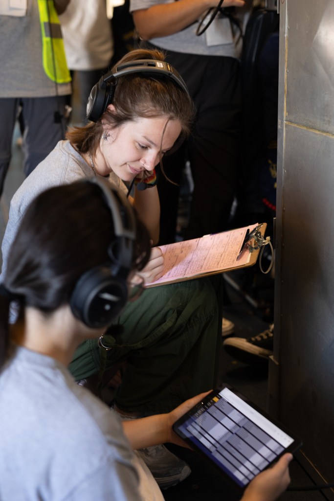 Two women wearing headphones and gray shirts sit on the floor, looking at a tablet with data and a clipboard.