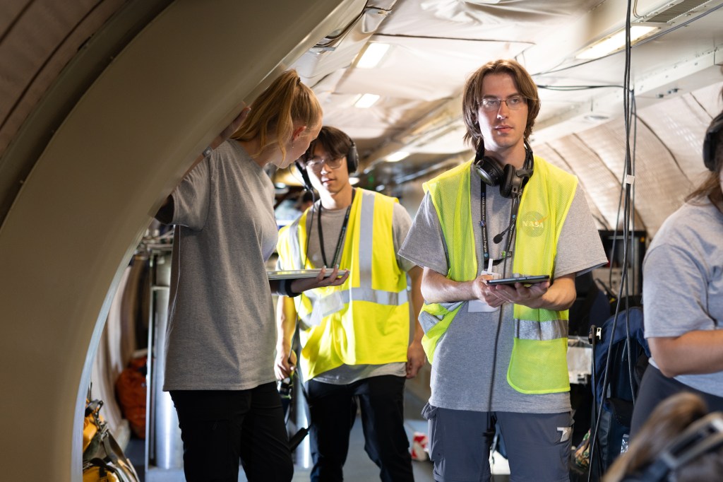 A student with brown hair, glasses, and a yellow high-viz vest writes on a clipboard.