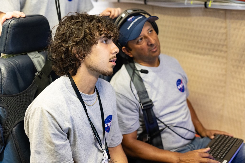 Two men in gray shirts with the NASA logo and headphones sit looking at a screen.