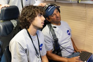 Two men in gray shirts with the NASA logo and headphones sit looking at a screen.
